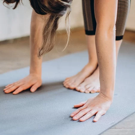 A person in a yoga pose with a close up of hands and bare feet on a grey-blue yoga mat.